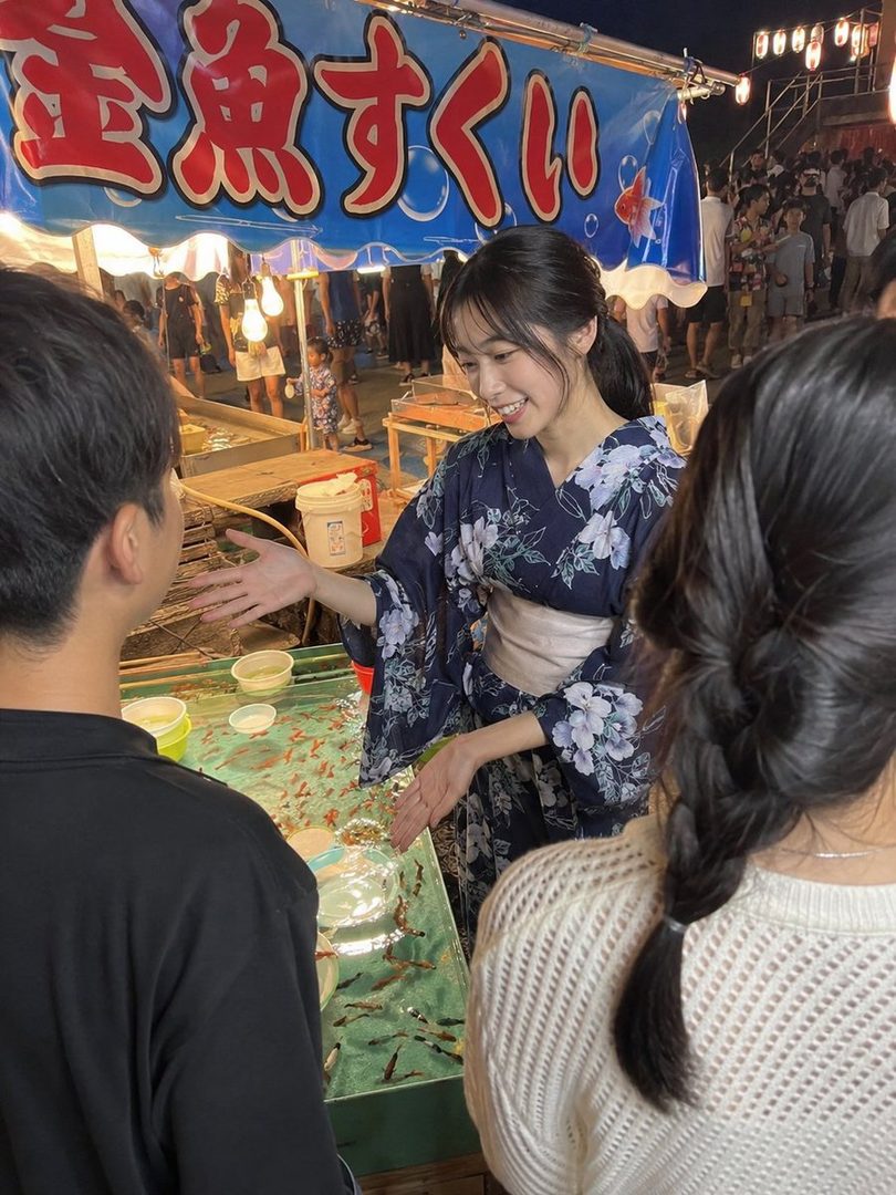 A young woman in a yukata smiles while explaining goldfish scooping at a night festival stall.