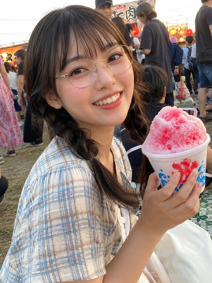 A smiling young woman with braided hair and glasses holds a cup of shaved ice at an outdoor event.