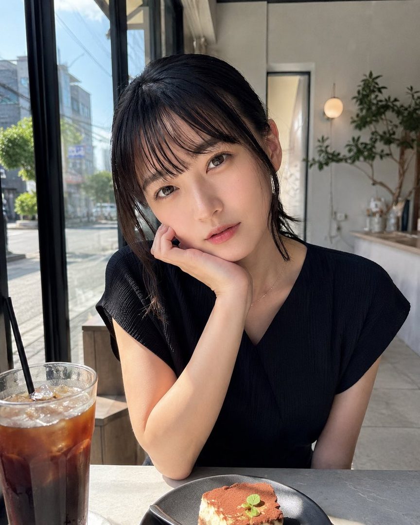 A young woman with black hair and bangs sits at a cafe table with iced coffee and dessert, looking thoughtfully at the camera.
