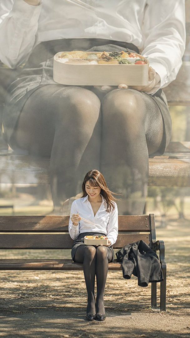 A smiling woman in a white blouse and black skirt sits on a park bench, eating from a bento box, with a large, blurred image of a person holding a bento box in the background.