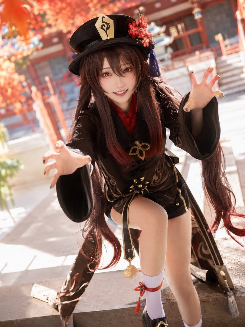 A cosplayer dressed as Hu Tao from Genshin Impact poses outdoors with a playful expression, wearing a black and red outfit with a matching hat.