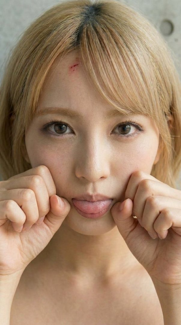 A playful portrait of a woman with blonde hair, sticking out her tongue and pulling her cheeks, with a small scratch on her forehead.