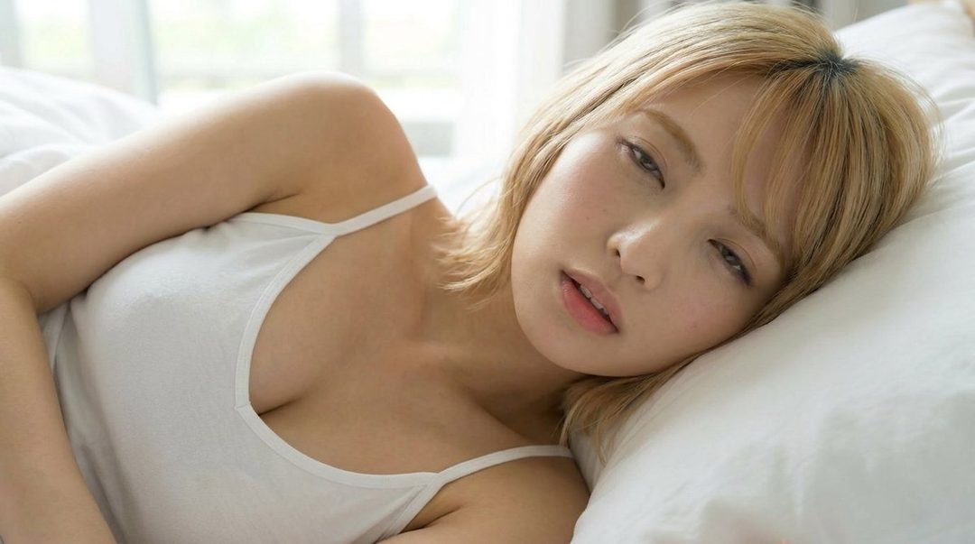 A blonde woman in a white tank top lies on a white pillow, looking dreamily at the camera.