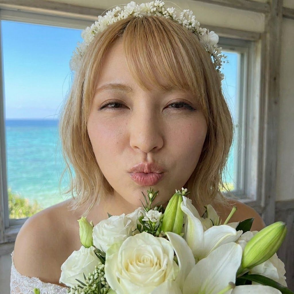 A blonde woman wearing a white floral crown and dress, holding a white bouquet, pouts her lips for the camera with a blue ocean in the background.