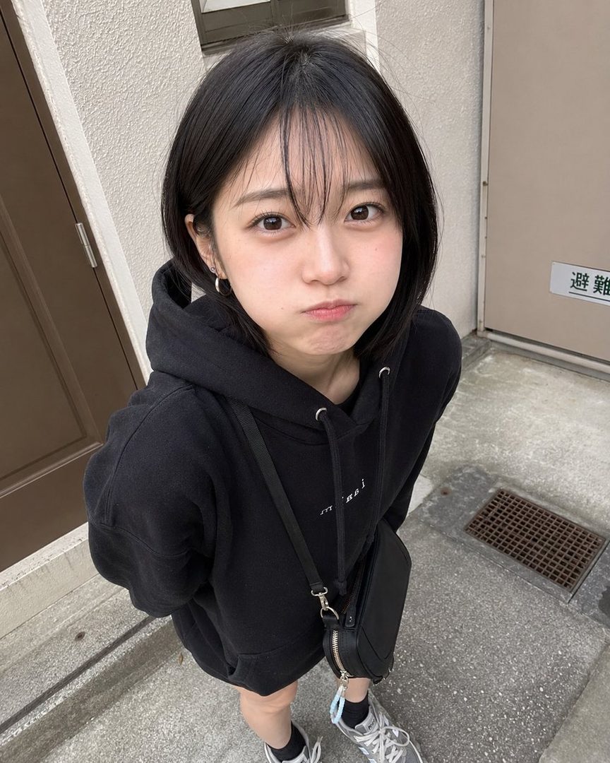 A young woman with short black hair and bangs, wearing a black hoodie, pouts at the camera outdoors.