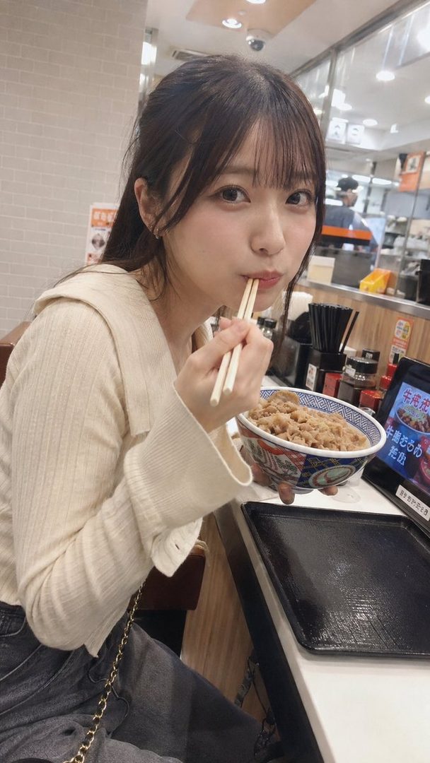 A young woman with brown hair and bangs is eating a bowl of gyudon with chopsticks in an indoor setting.