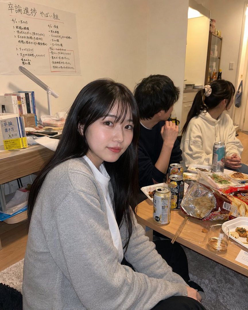 A young woman with long black hair and a grey cardigan smiles gently at a casual home gathering with friends, surrounded by snacks and drinks.