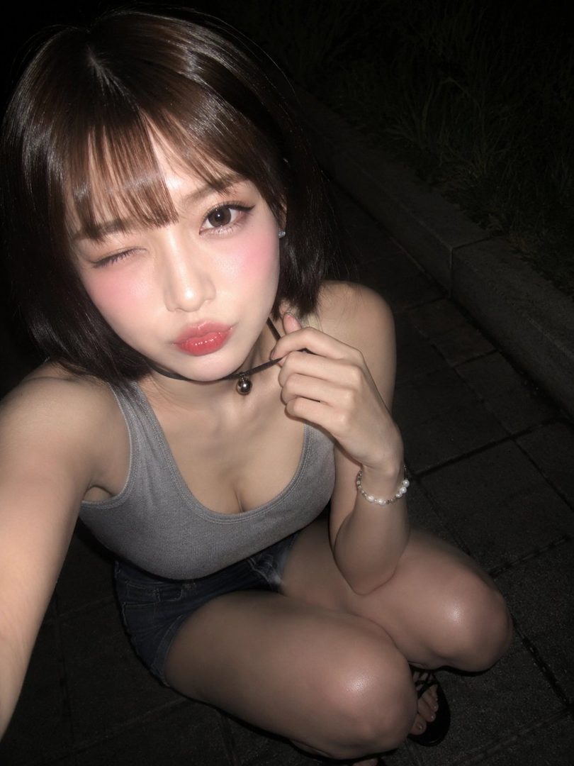 A young East Asian woman winks at the camera in a close-up night selfie, wearing a gray tank top and dark shorts.