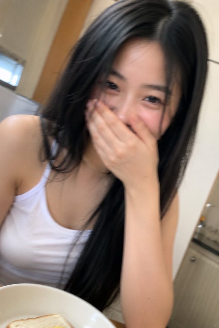 A candid shot of a young East Asian woman with long black hair, covering her mouth with her hand while smiling, wearing a white tank top.