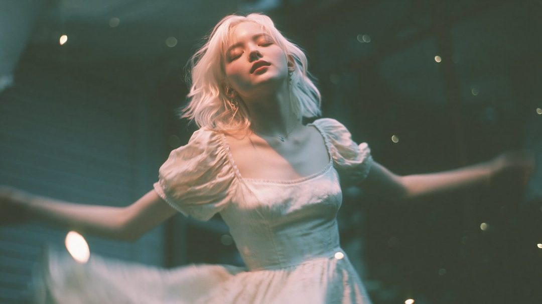 A dreamy portrait of a blonde woman in a white dress with her eyes closed, surrounded by soft lights.