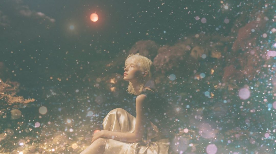 A blonde woman in a dress looks up at a red moon in a dreamy, star-filled night sky.