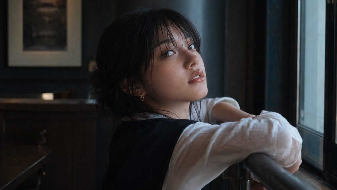 A young woman with short black hair and bangs looks over her shoulder while leaning on a window sill indoors.