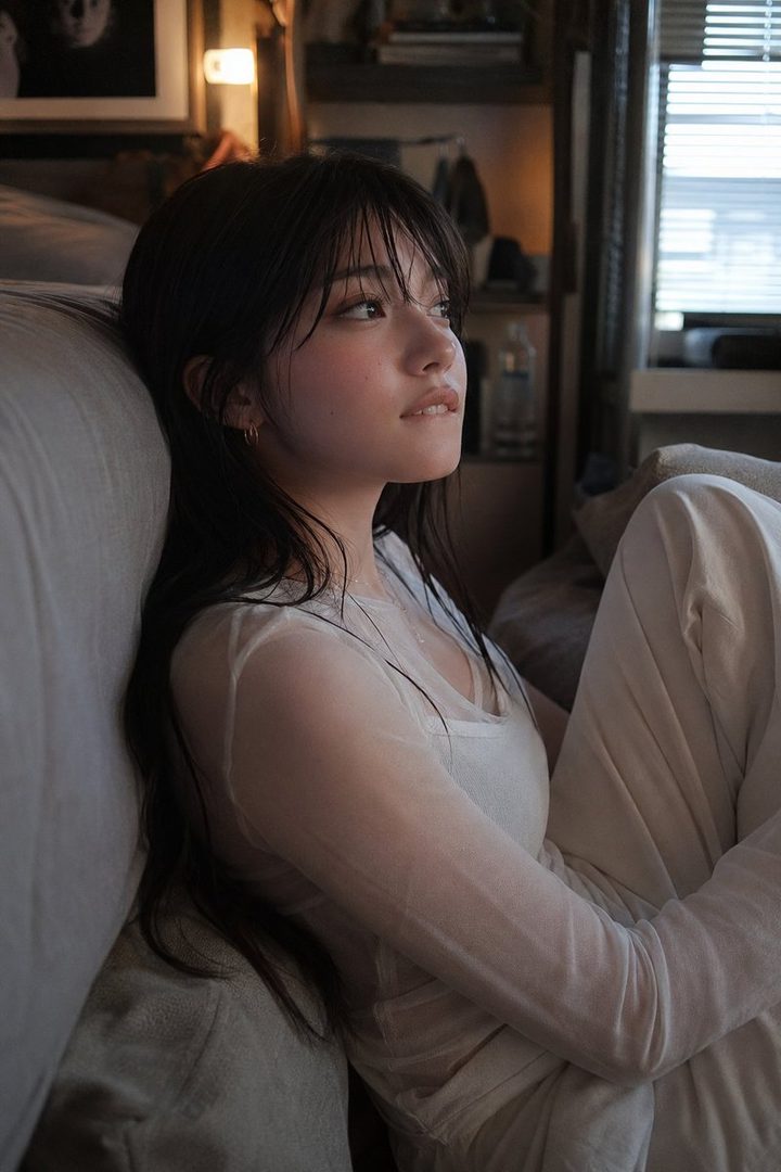 A young woman with long dark hair and bangs is sitting indoors, looking thoughtfully to the side.