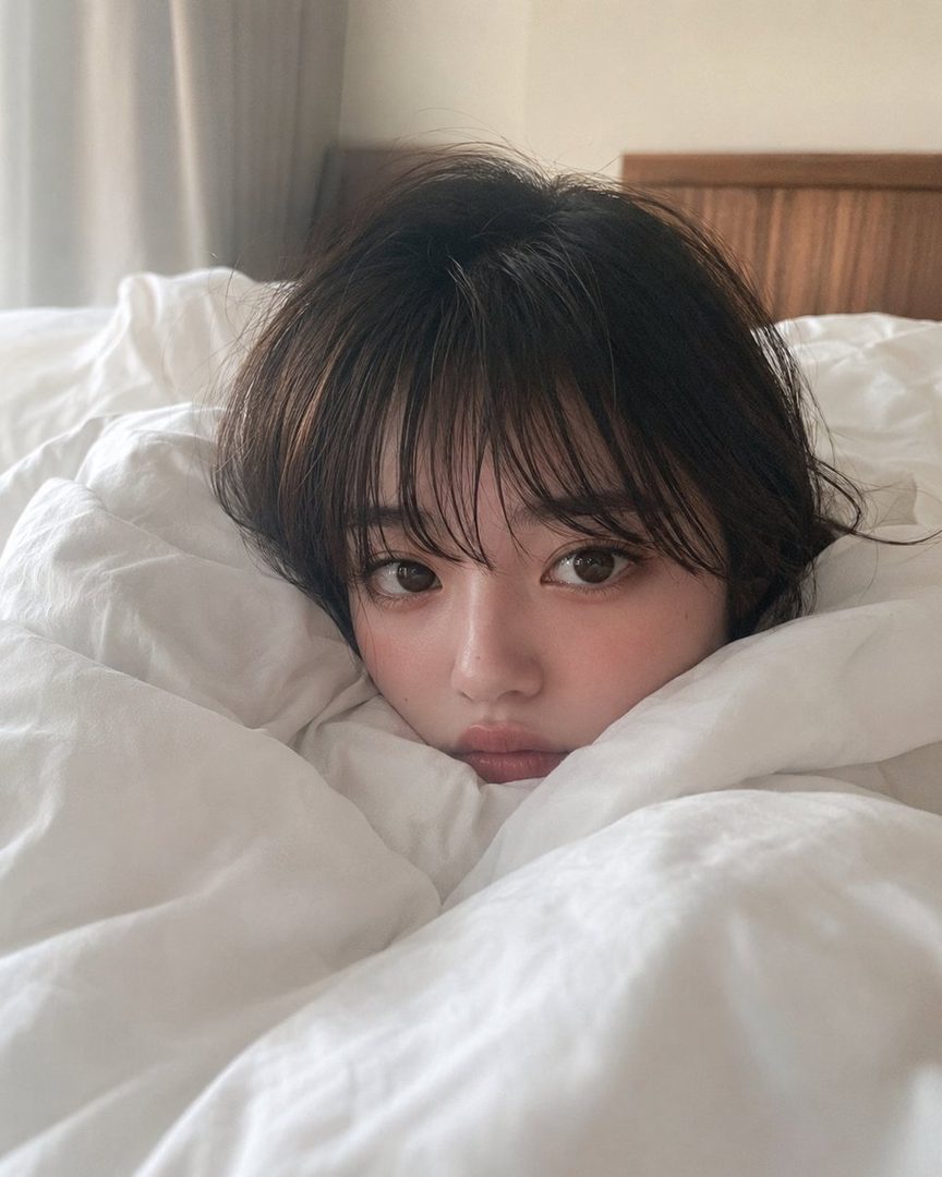 A cute woman with short brown hair and bangs peeks out from under a white duvet in a bedroom.