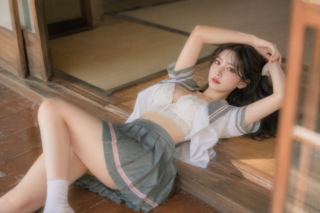 A young woman in a school uniform top and short skirt is lying on a wooden floor indoors, looking at the camera.