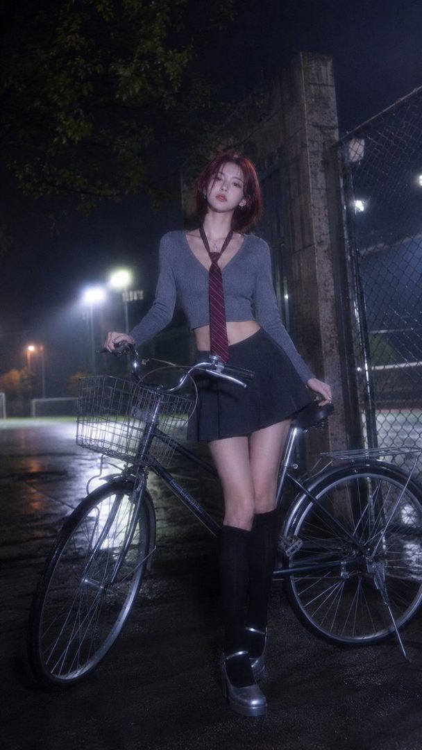 A young woman with short red hair and a tie stands next to a bicycle at night.