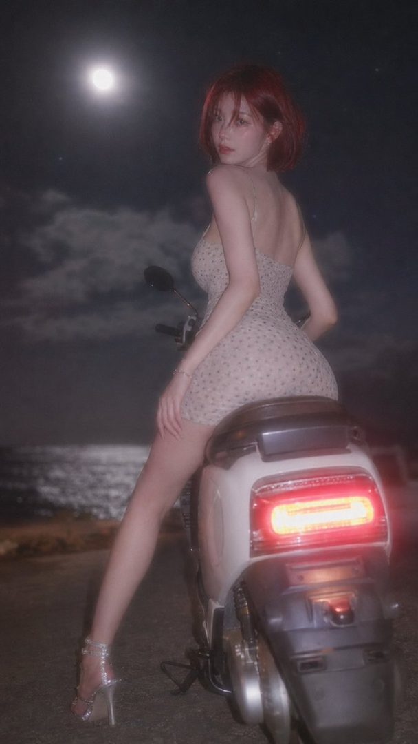 A woman with red hair in a short dress sits on a scooter at night with the moon in the background.
