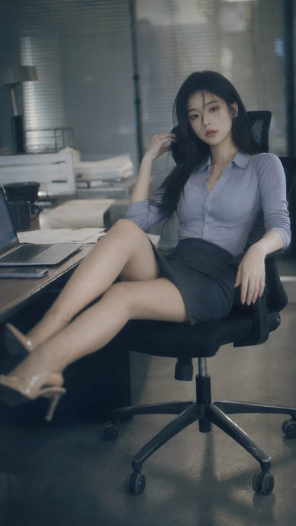 A young East Asian woman with long black hair sits in an office chair, wearing a light blue blouse and a dark skirt, with her legs propped on a desk.