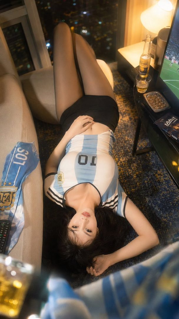 A woman wearing an Argentina soccer jersey and black shorts lies on the floor in an indoor setting at night, with city lights visible outside a window.