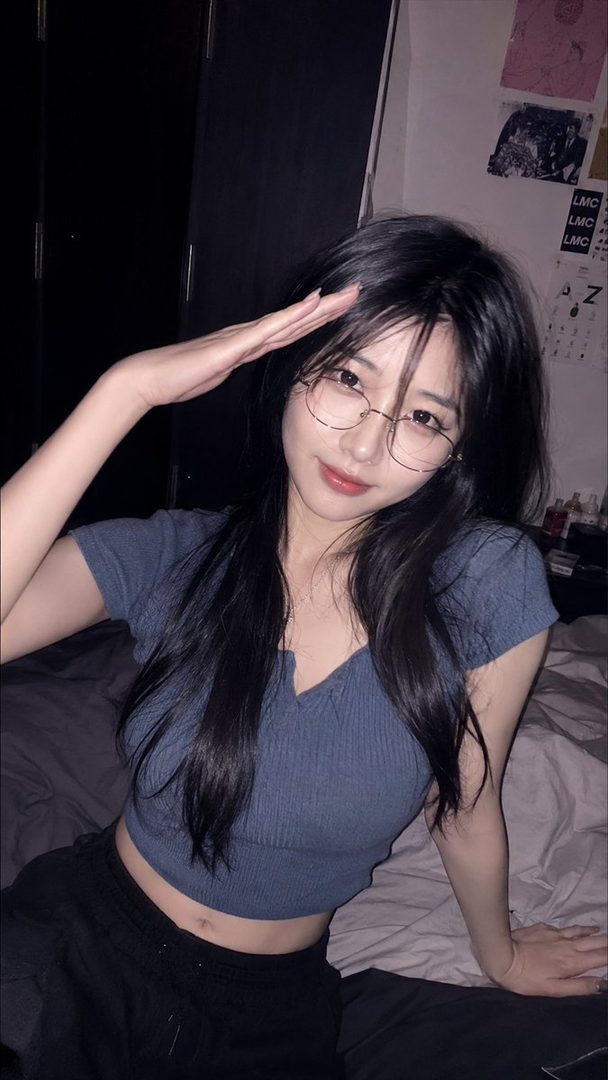 A young woman with long black hair and glasses poses playfully in a dim bedroom, wearing a blue crop top.