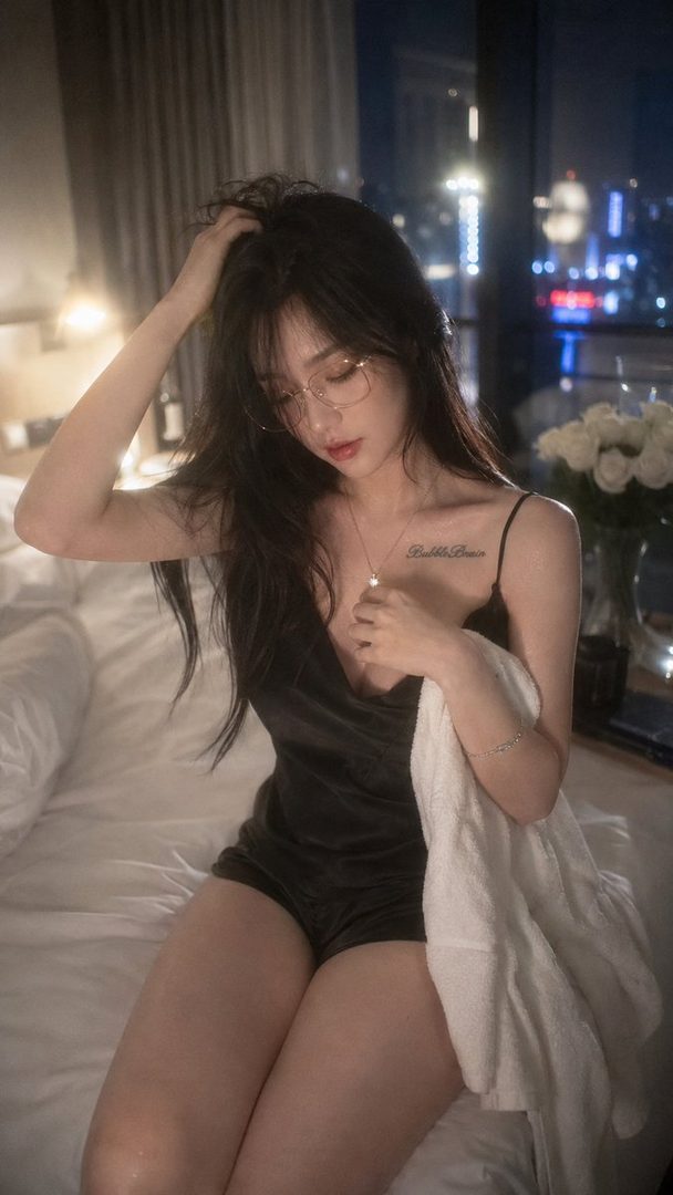 A woman with long black hair and glasses sits on a bed in a hotel room at night, wearing a black camisole and shorts.