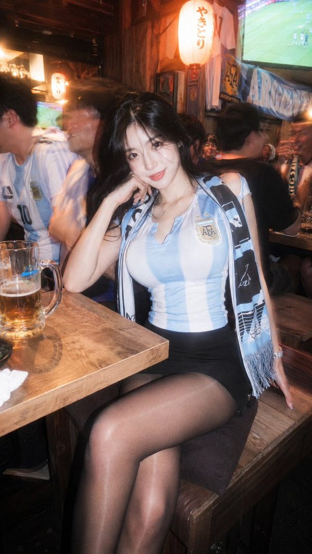 A young woman in an Argentina jersey and scarf sits at a table in a lively izakaya during a World Cup match.