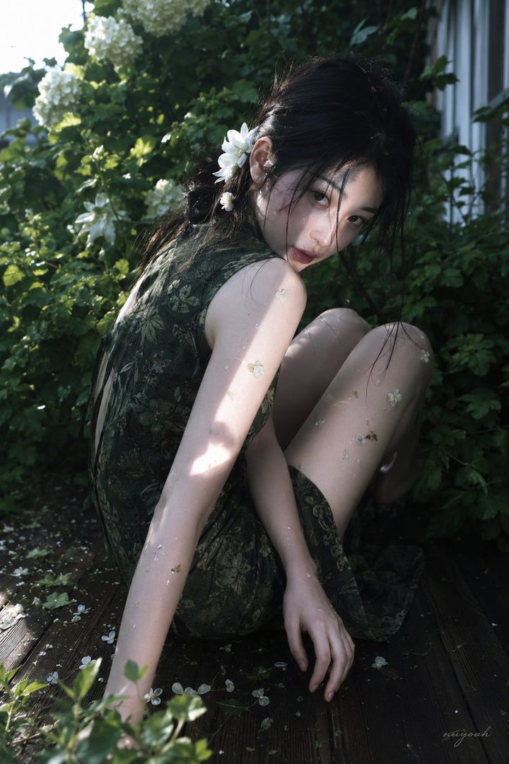 A young woman with black hair and a green dress sits among green foliage and white flowers, looking back at the camera with a dreamy expression.