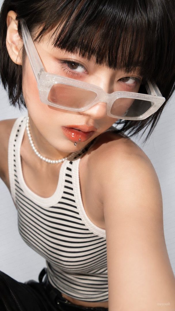 A close-up portrait of a young woman with short black hair and bangs, wearing a striped tank top, a pearl necklace, and white sunglasses, looking directly at the camera.