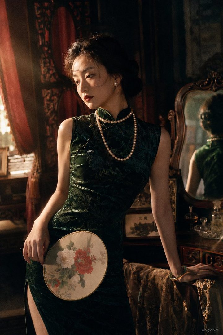 A woman in a dark green cheongsam with a pearl necklace holds a traditional fan, posing elegantly indoors with a vintage aesthetic.