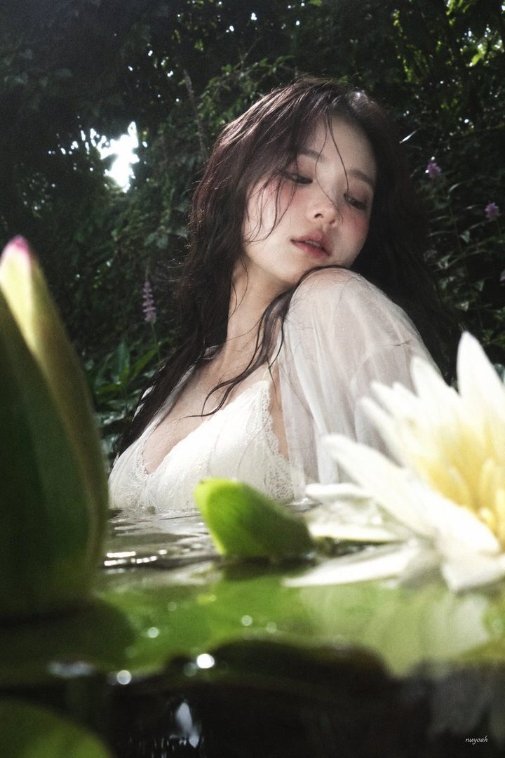 A dreamy portrait of a woman with wet brown hair and a white lace top, partially submerged in water with water lilies and green foliage.