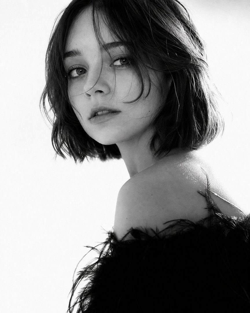 A black and white portrait of a woman with short dark hair and bangs, wearing a feathered top, looking directly at the camera.