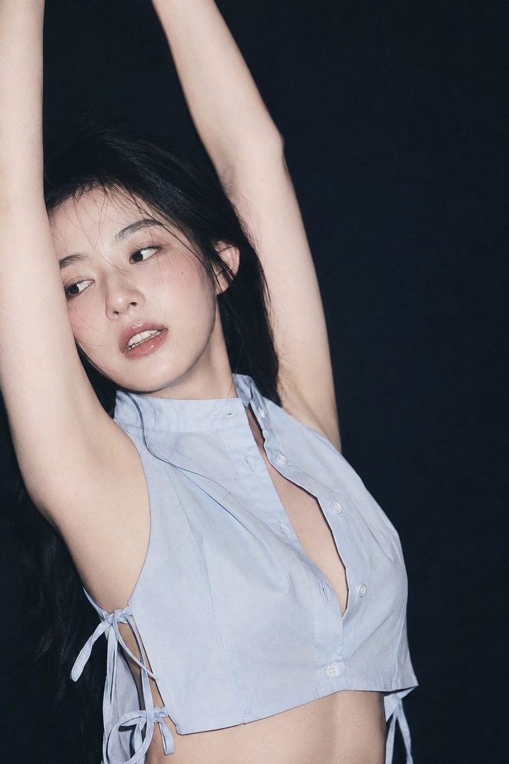 A beautiful East Asian woman with long black hair poses in a light blue sleeveless top with her arms raised against a dark background.