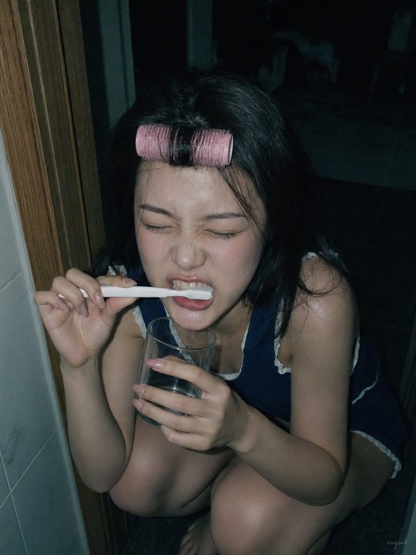 A young East Asian woman with black hair and a pink hair roller brushes her teeth while holding a glass of water, captured in a Y2K-style indoor portrait.