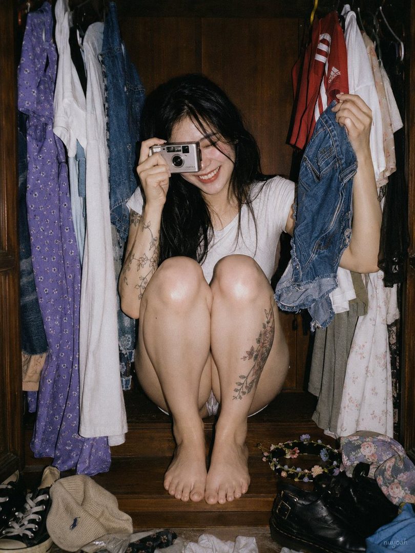A young woman with long black hair and tattoos, wearing a white t-shirt, sits in a closet holding a camera and smiling playfully.