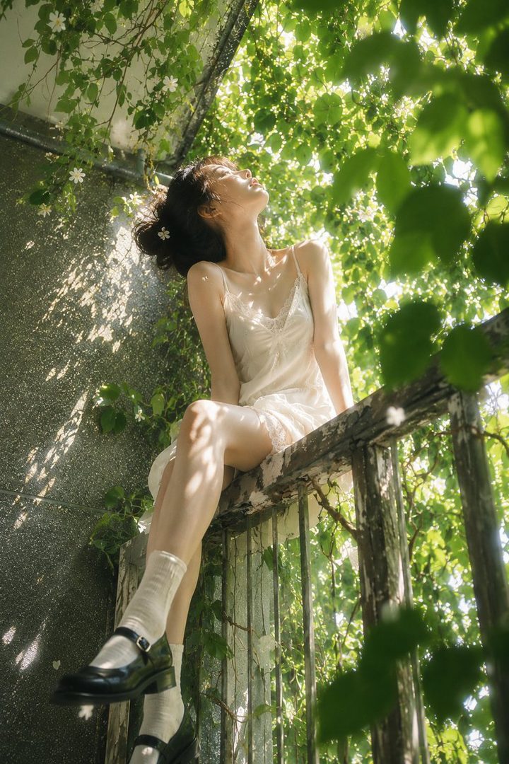 A woman in a white slip dress sits on a wooden railing surrounded by lush green foliage, looking up at the sunlight filtering through the leaves.