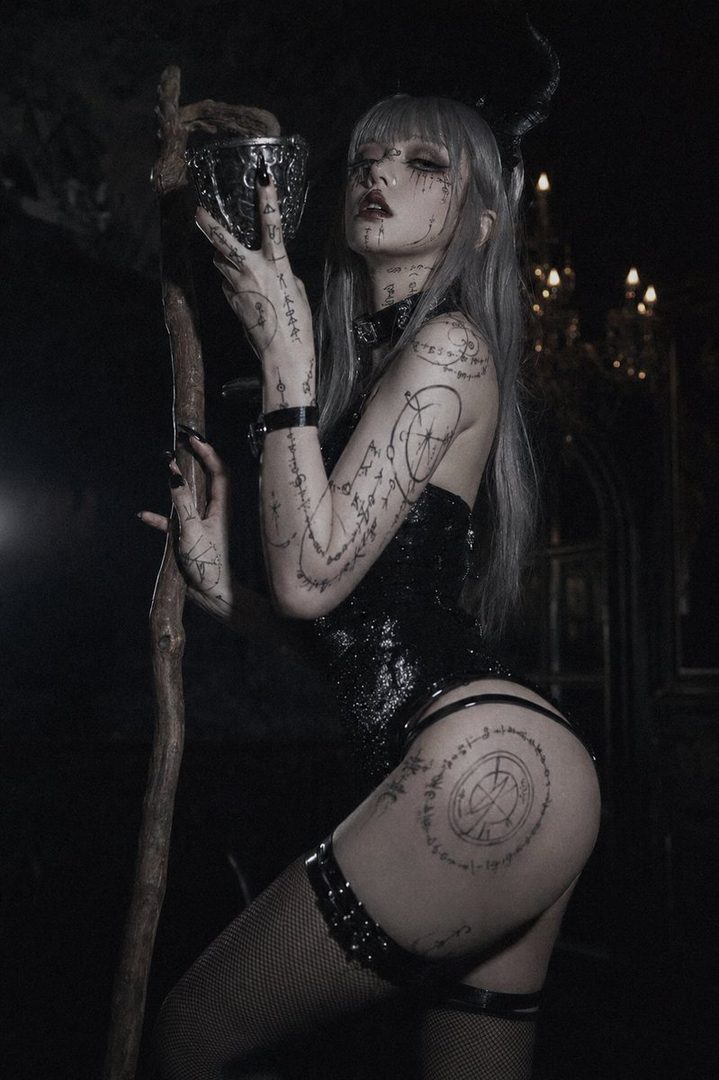 A woman with silver hair and dark makeup, adorned with body art, holds a goblet and staff in a dark, gothic setting.