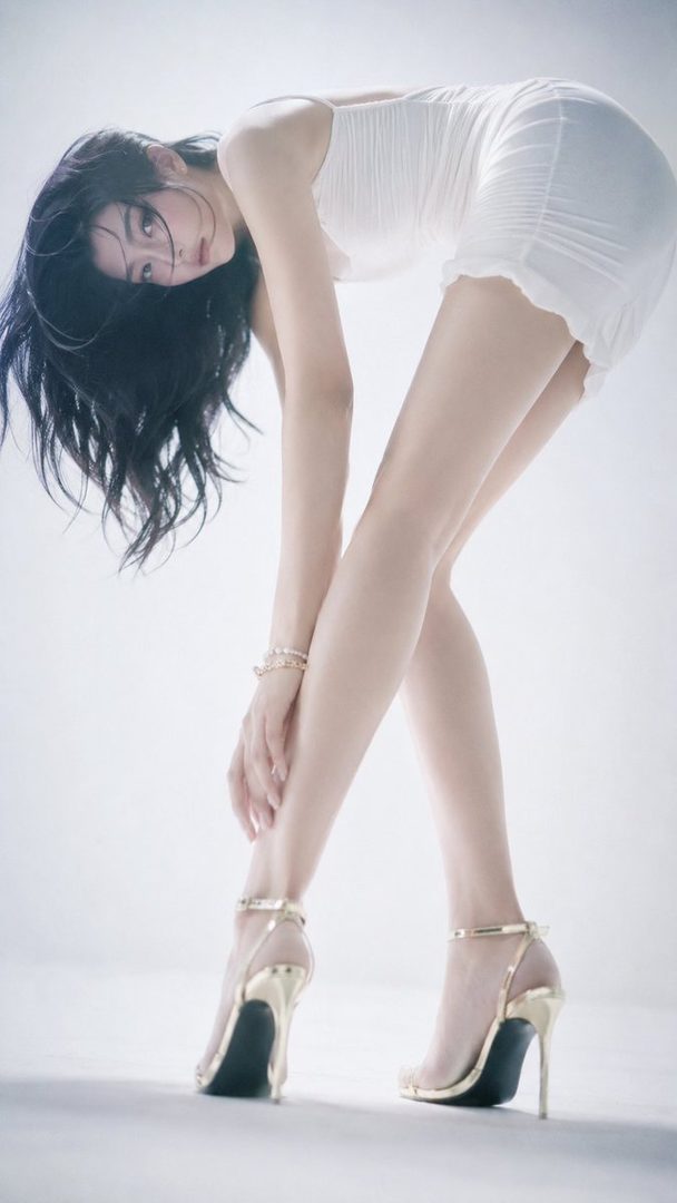 A woman with long black hair in a white dress and gold high heels bends over in a studio setting.