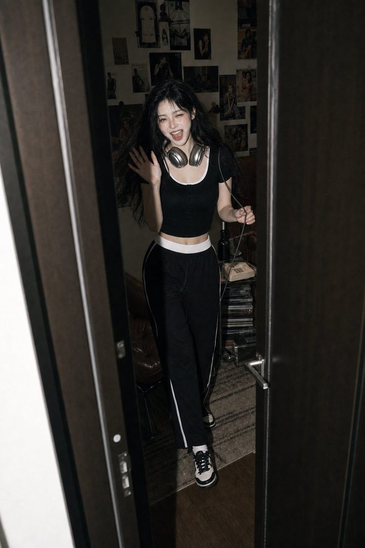 A playful woman with long black hair and headphones around her neck, wearing a black crop top and track pants, waves from a doorway in a dimly lit room.