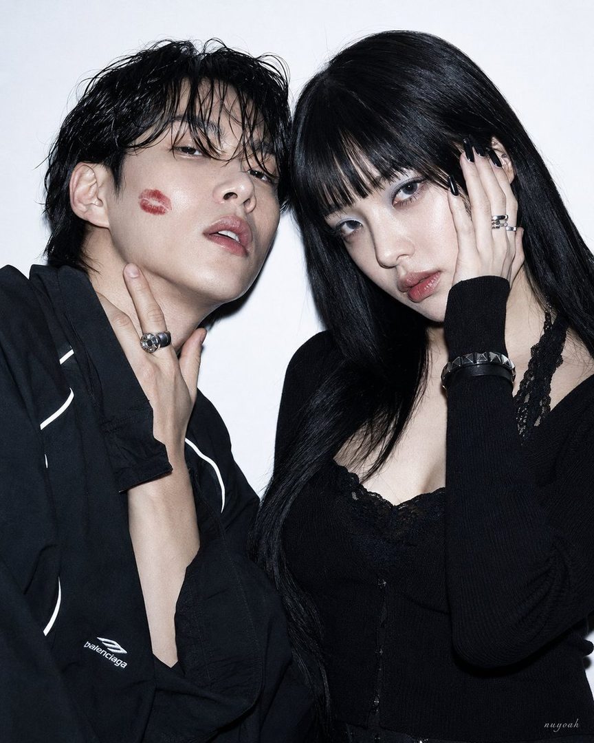 A man and a woman with black hair pose closely in a studio, both wearing black outfits and rings, with a red lipstick mark on the man's cheek.