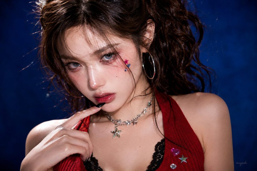 A close-up studio portrait of a woman with curly brown hair, wearing a red halter top and black lace, with star stickers on her cheek and a star necklace.