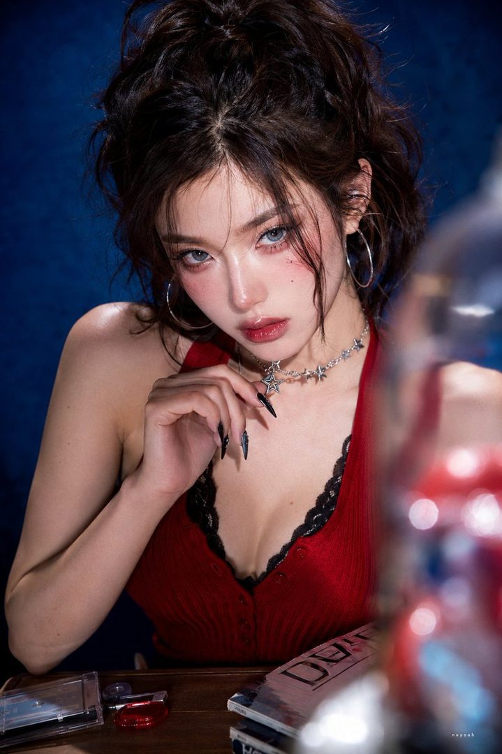A woman with dark hair and a red top poses in a studio with a blue background, wearing a star necklace and large hoop earrings.