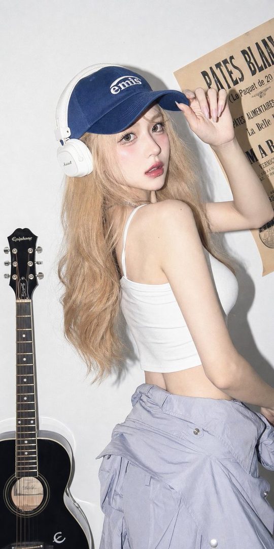 A young woman with blonde hair and a blue cap poses indoors with a guitar and a vintage poster in the background.