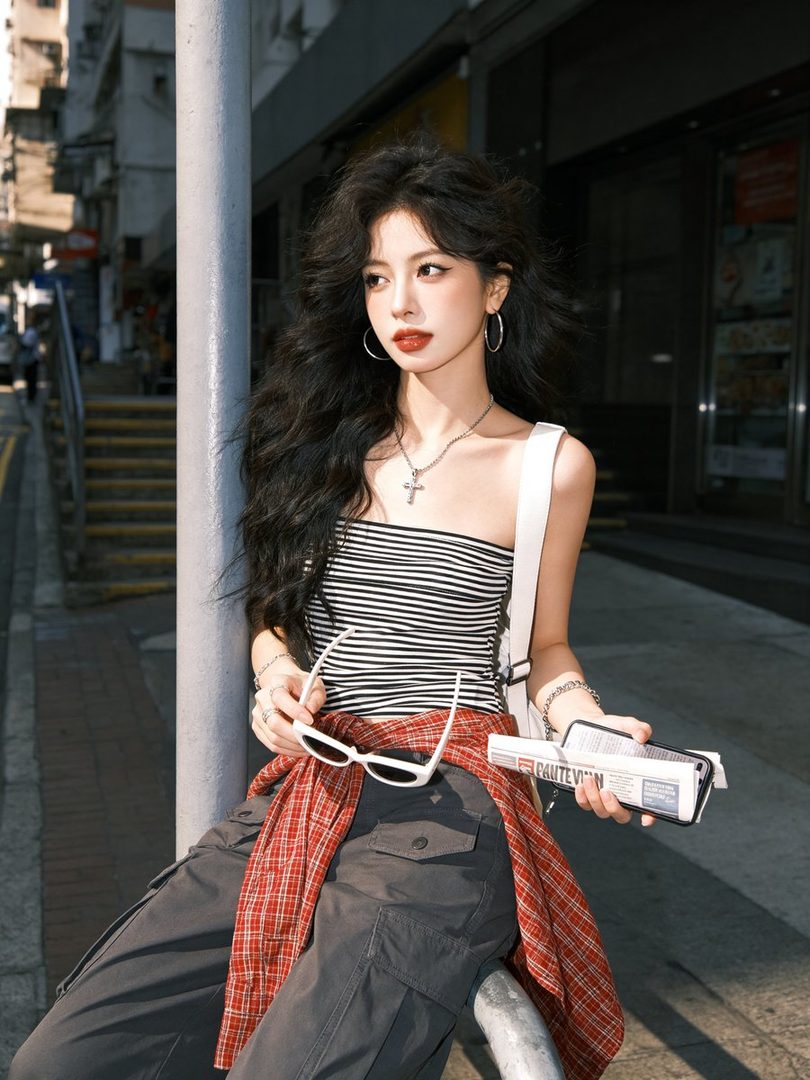 A young woman with long black hair and a striped top poses on a city street, holding sunglasses and a newspaper.