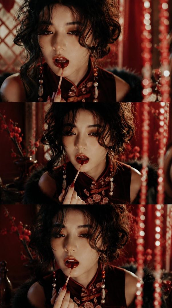 A woman with dark curly hair and a red traditional dress applies red lipstick, with a seductive expression.