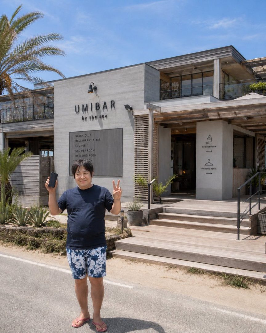 A man in a dark blue t-shirt and floral shorts stands in front of a building named UMIBAR by the sea, holding a phone and making a peace sign.