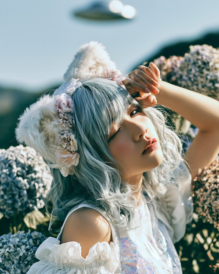 A woman with silver hair and cat ears headband poses outdoors with hydrangeas and a blurred UFO in the background.
