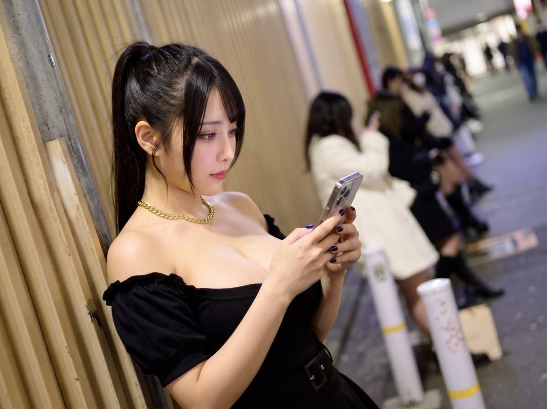 A woman with black hair in a ponytail and an off-shoulder black dress is looking at her phone on a city street at night.