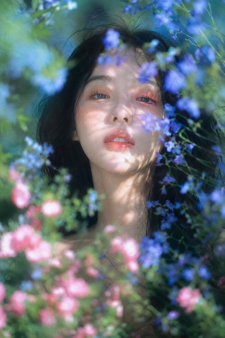 A dreamy portrait of a woman with long black hair, surrounded by pink and blue flowers in a garden, with dappled sunlight on her face.