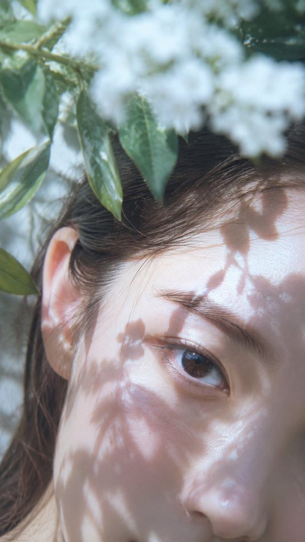 A close-up portrait of a woman's face with dappled sunlight and green leaves in the foreground.