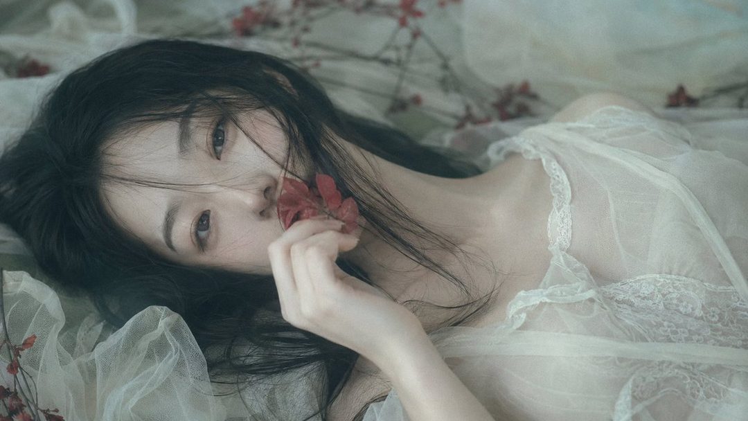 A woman with long black hair lies on a bed, holding a red flower to her lips, with a dreamy and serene expression.